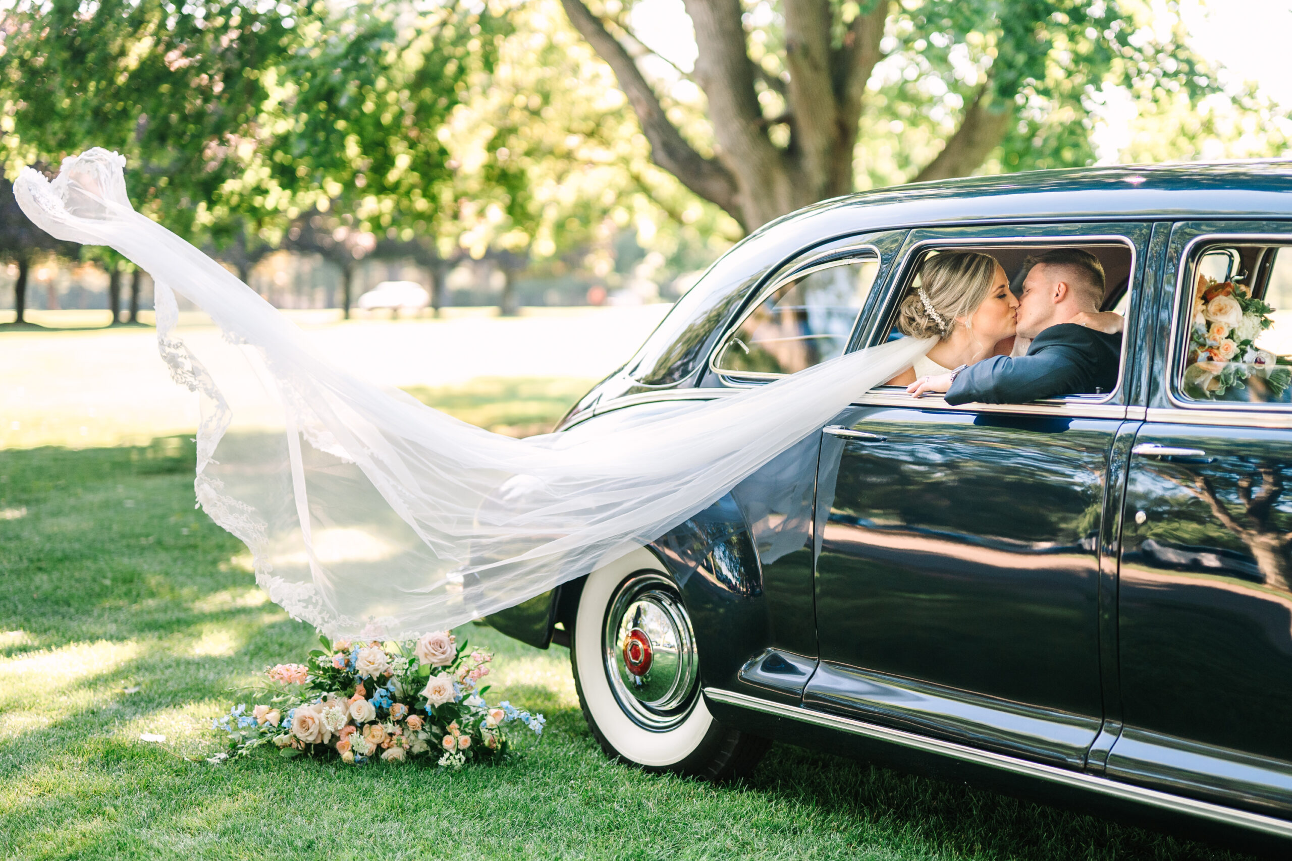 A couple kisses int heir vintage getaway car as the veil flies out the window