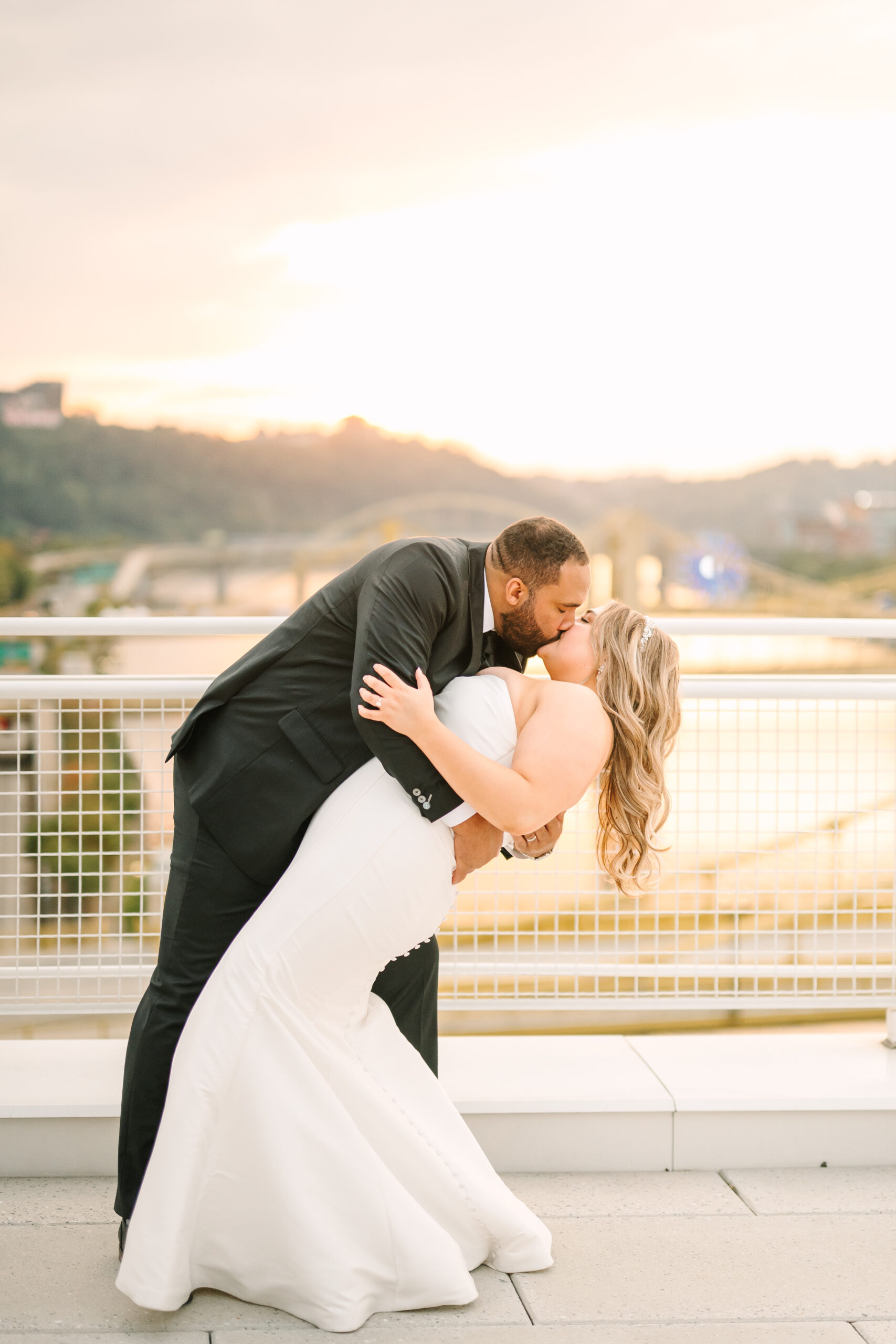 A newlywed couple shares a kiss on the rooftop of the David L. Lawrence Convention Center, with the Pittsburgh skyline in the background. The bride, in a beautiful white gown, wraps her arms around her partner, who is dressed in a classic black suit. The city’s tall buildings and a golden sky create a stunning backdrop, capturing the romantic and celebratory moment.