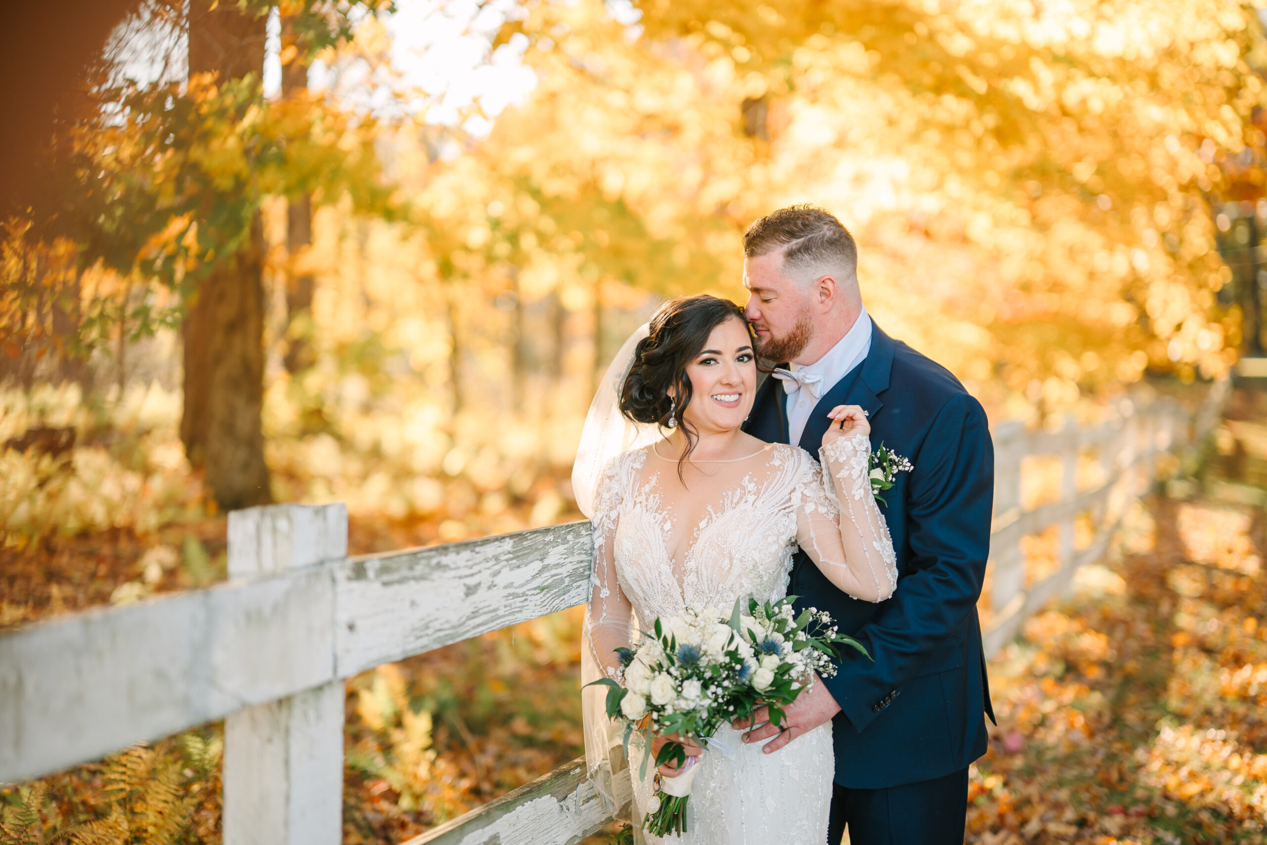 A bride and groom stand together under a canopy of vibrant fall foliage, with golden leaves scattered on the ground and warm sunlight filtering through the trees, creating a romantic and timeless autumn wedding scene.